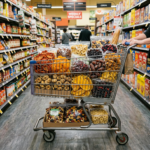 Shopping cart filled with groceries including milk, bananas, cereal, bread, vegetables, and laundry detergent